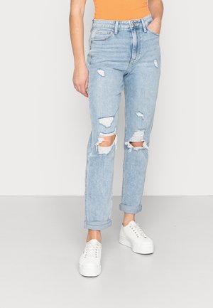 Person iført lyseblå revnede jeans med opsmøg, hvide plateausneakers og en orange ærmeløs top, stående mod en ensfarvet baggrund.