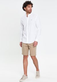 Mann trägt weißes langärmliges Hemd mit Knöpfen, beige Shorts und hellbeige Sneakers, steht mit zur Seite gedrehtem Kopf vor weißem Hintergrund.