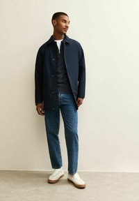 Giacca leggera blu navy con zip frontale, due tasche e chiusure a scatto, abbinata a un maglione scuro, jeans blu e sneakers bianche.