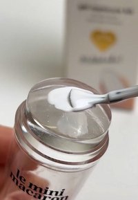 Przezroczysty słoik z przezroczystą pokrywką zawiera gładki biały produkt, nakładany małą szpatułką, o zaokrąglonym kształcie.