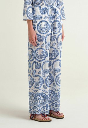 Pantaloni bianchi a gamba larga con stampa paisley blu indossati con sandali rossi e neri con motivi su una persona in piedi.