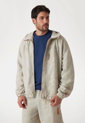 Svetlo bež zip-up jakna z elastiko na rokavih, ki vsebuje nevsiljiv vzorec, logo na prsih in manjšo zadrgo žep. V kombinaciji z modro majico.