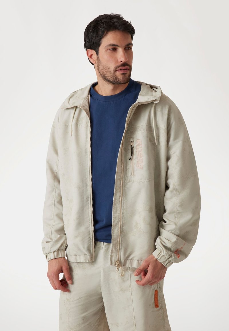 Light beige zip-up jacket with elastic cuffs, featuring a subtle pattern, logo on the chest, and a small zippered pocket. Paired with a blue shirt.
