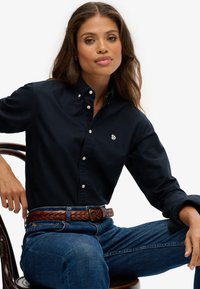 Camisa de botón en color navy hecha de algodón, con un sutil logo bordado. Combinada con jeans de mezclilla azul y un cinturón de cuero trenzado marrón.