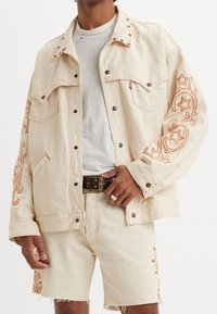 Béžová džínová bunda s dekorativním oranžovým vyšíváním a límečkem ozdobeným cvoky, doplněná o odpovídající šortky a černý opasek s cvoky.