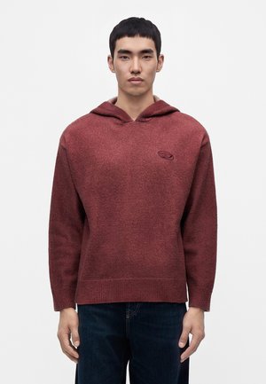 Jeune homme portant un pull en maille à capuche bordeaux et un jean foncé, debout devant un fond blanc uni.