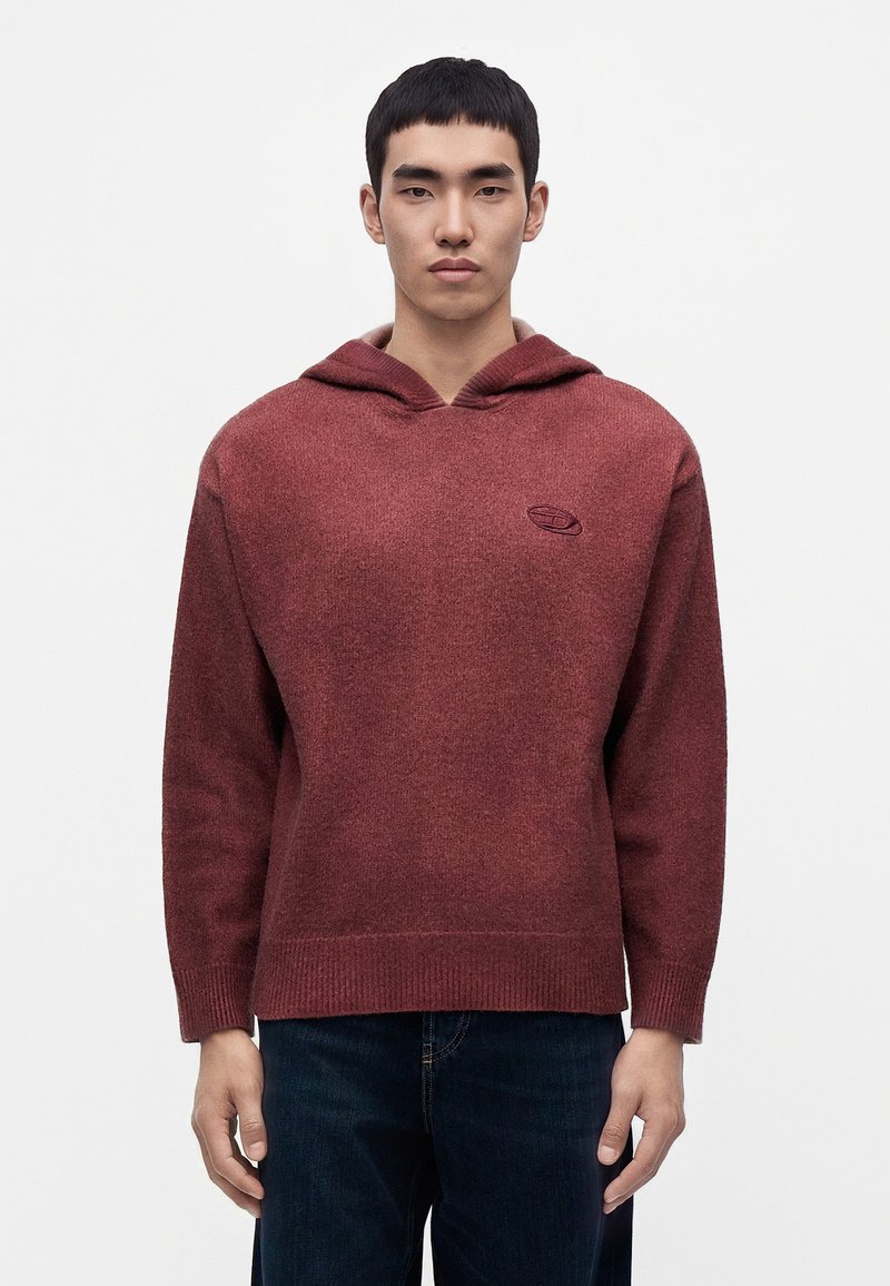 Jeune homme portant un pull en maille à capuche bordeaux et un jean foncé, debout devant un fond blanc uni.
