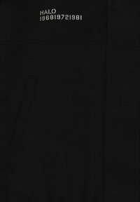 Tissu noir avec une poche verticale cousue et un texte blanc "HALO 196819721981" imprimé au-dessus de la poche.