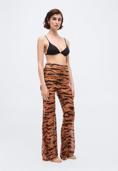 Γυναίκα που στέκεται ξυπόλυτη, φορώντας μαύρο σουτιέν και καφέ παντελόνι με ρίγες τίγρης, σε φαρδιά γραμμή, μπροστά σε λευκό φόντο.