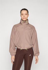 Giacca cropped color marrone chiaro realizzata in materiale morbido, con colletto alto, polsini elasticizzati e cordini sul bordo in un colore a contrasto.