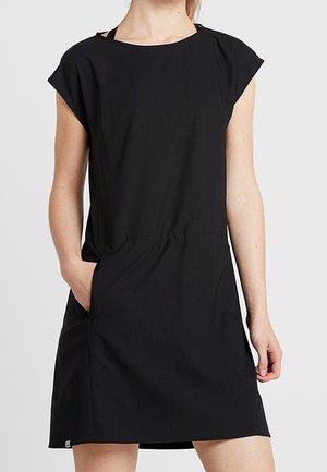 Femme portant une courte robe noire sans manches avec un col montant et des poches latérales, debout devant un fond clair uni.