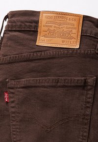 Jean en denim marron avec un patch en cuir portant le logo « Levi Strauss & Co. » orné d'un dessin de deux chevaux et une couture surélevée. Onglet rouge sur la poche.