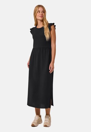 Femme aux longs cheveux blonds portant une robe midi noire sans manches avec des épaules volantées et des baskets beige clair, debout et regardant sur le côté.