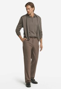 Herrens outfit består av en grå långärmad pikétröja under en matchande zip-up cardigan, kompletterad med bruna rutiga byxor och svarta skor.