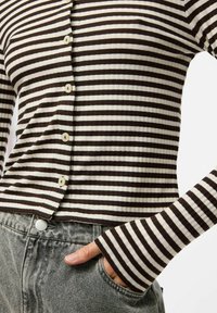 Halenka s dlouhým rukávem, žebrovaná, s knoflíky a střídavými tmavě hnědými a krémovými pruhy; má kulaté knoflíky a těsný střih. Šedé džíny s vysokým pasem.