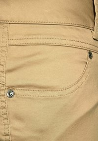 Close-up van een beige broekzak met zichtbare stiksels en twee metalen klinknagels op de stof.