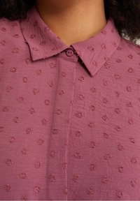 Chemise couleur pin avec un tissu texturé par des motifs floraux en relief, un col classique et une fermeture à boutons au niveau du cou.