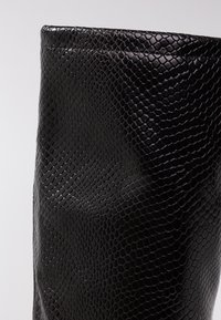 Caño de bota negra con un patrón de piel de serpiente brillante y texturizado y costuras visibles a lo largo del borde superior sobre un fondo neutro.