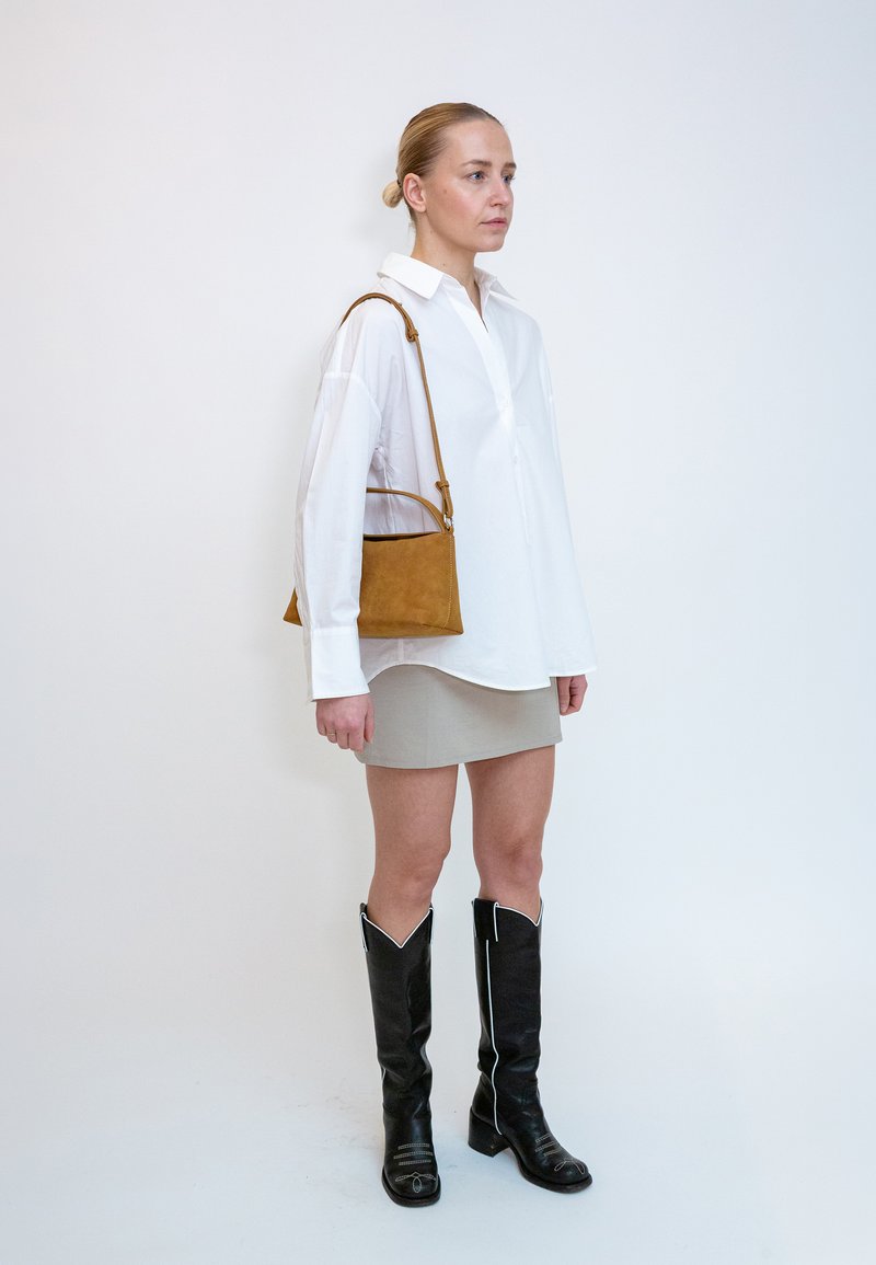 Femme debout portant une chemise blanche oversize, une mini-jupe beige, des bottes noires montantes jusqu'aux genoux et un sac à bandoulière marron.