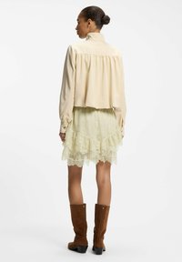 Mujer con el pelo recogido en un moño, que lleva una blusa de manga larga color beige claro, falda con volantes de encaje crema y botas marrones de ante hasta la rodilla, de espaldas.