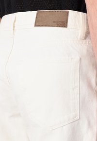 Pantalones de denim blancos con un parche de cuero marrón etiquetado "SELECTED HOMME", con un diseño clásico de cinco bolsillos y detalles de costura sutiles.