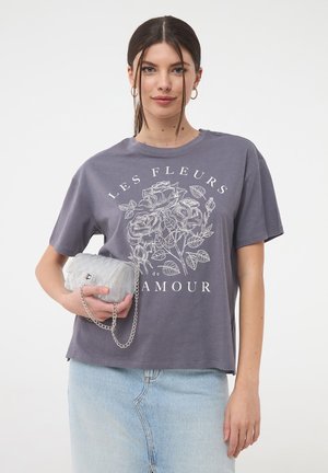 T-shirt grigia con stampa floreale bianca, maniche corte e vestibilità rilassata. Tiene in mano una piccola borsa argentata con tracolla a catena.