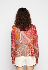 Camisa de manga longa vermelha e laranja com padrão paisley, textura leve e punhos com botão, combinada com calções creme.