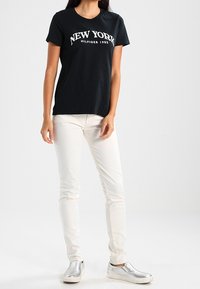 Černé tričko s nápisem "NEW YORK" tištěným bílou barvou, doplněné o úzké bílé kalhoty a stříbrné slip-on boty. Hladká bavlněná textura.