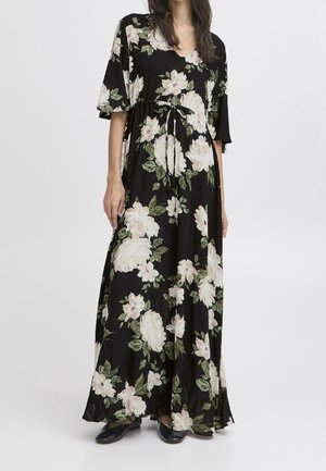 Femme portant une robe noire longue jusqu'aux chevilles avec un grand imprimé floral blanc et vert et des manches courtes, associée à des chaussures noires.