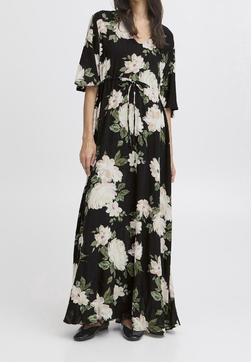 Femme portant une robe noire longue jusqu'aux chevilles avec un grand imprimé floral blanc et vert et des manches courtes, associée à des chaussures noires.