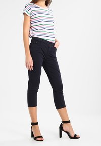 Kvinna klädd i randig kortärmad topp, avklippta svarta byxor och svarta höga sandaler med ankelsläp, står med en hand i fickan.