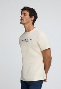 Krämfärgad bomullst-shirt med korta ärmar. Har texten "POLO CLUB" i svart och rött över bröstet. Normal passform.