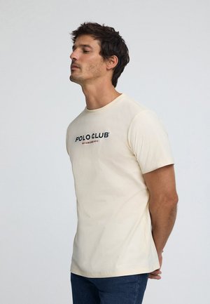 Kremfarvet bomuld T-shirt med korte ærmer. Har teksten "POLO CLUB" i sort og rød tværs over brystet. Normal pasform.