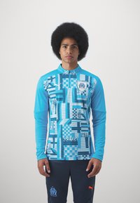 Maglietta sportiva azzurra a maniche lunghe con vari motivi geometrici e colletto con zip, indossata con pantaloni navy e un logo arancione.