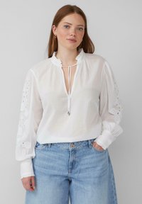 Blouse blanche avec un col à volants et des liens ajustables. Présente des détails brodés sur les manches. Portée avec un jean taille haute bleu clair.