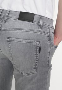 Los jeans de mezclilla gris presentan un corte ajustado, bolsillos traseros y una etiqueta de cuero negro en la cinturilla. La tela tiene una textura suave.
