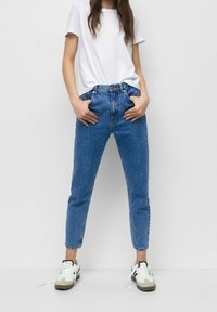 Jean en denim bleu taille haute avec une coupe droite, cinq poches et une fermeture à bouton. Associé à un t-shirt blanc uni et des baskets.