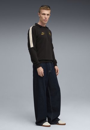 Sort langærmet polo med guld logo, mønstrede skulderstriber, parret med blå vide jeans og hvide sneakers.
