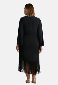 Černé šaty s dlouhými rukávy, zadním otvorem ve tvaru klíče a detailem třásní na spodním lemu; vyrobené z hladké látky s volným střihem.