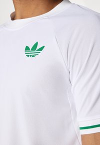 Valge sportlik särk, millel on rinnal roheline Adidas'i logo, võrkmaterjal ja roheline triibuline aktsent varrukatel.