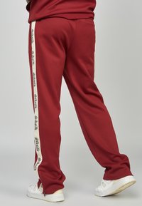 Bordeauxrote Jogginghose mit seitlichem Reißverschluss, weißem Akzentstreifen mit "Siksilk"-Logo. Hergestellt aus weichem Stoff, entspannte Passform, elastischer Bund.