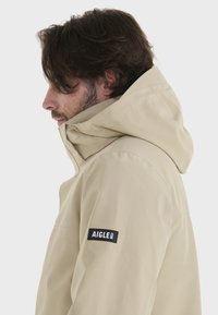 Beige vandtæt jakke med hætte, der har en justerbar krave, sidelommer og et sort mærketikette på venstre ærme.