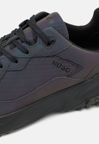 Tummansiniset tennarit, joissa on sileä, matta pinta, teksturoitu yksityiskohta, kumipohja, mustat nauhat ja "HUGO" -brändäys sivussa.