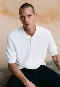 Homme aux cheveux courts et tatouages portant un polo blanc à manches courtes et un pantalon foncé, assis contre un mur beige.
