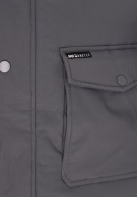 Grijze jas met een gladde textuur, voorzien van een zak met een logo, drukknopen en zichtbare stiksel details.