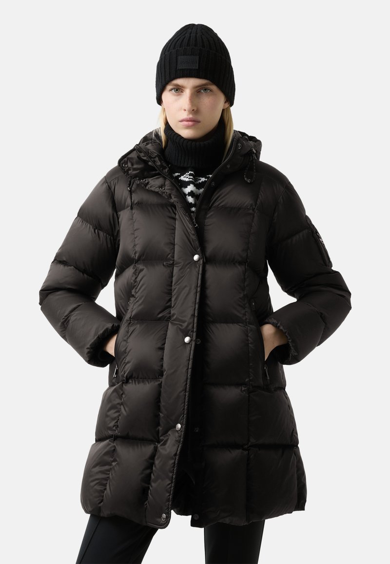Černý péřový kabát se zapínáním na zip, kovovými druky a kapucí. Nositelný přes černobílý pletený svetr a v kombinaci s černou čepicí.