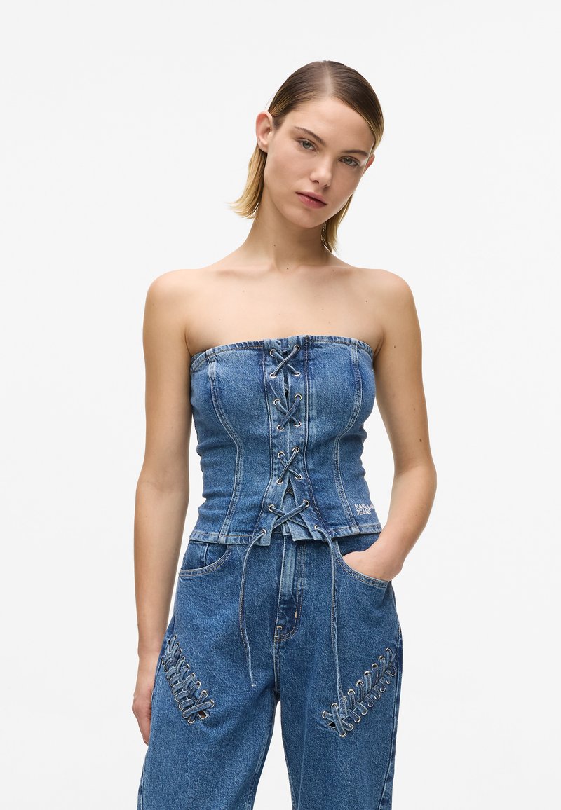 Νεαρή γυναίκα με κοντά μαλλιά, που φοράει μπλε Denim τοπ χωρίς τιράντες με κορδόνια και τζιν παντελόνι με λεπτομέρειες κορδονιών, στέκεται με το ένα χέρι στην τσέπη.