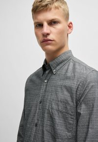 Camisa gris de botonadura con un sutil patrón texturizado, un bolsillo en el pecho y botones en contraste. Ajuste entallado y diseño de cuello curvado.