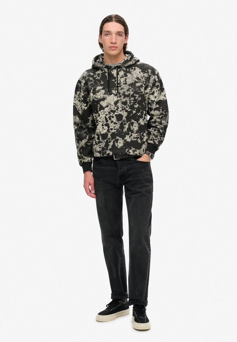 Czarna bluzka z kapturem z białym wzorem w plamy. Połączona z czarnymi dżinsami i czarnymi trampkami, charakteryzującymi się teksturowanym materiałem i luźnym krojem.