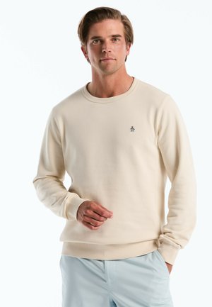 Jeune homme portant un sweat-shirt beige à col rond avec un petit logo de pingouin sur la poitrine et un pantalon bleu clair, debout devant un fond blanc.